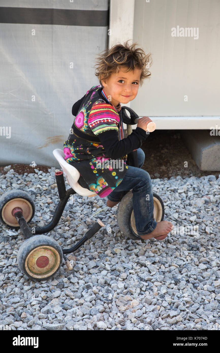 Souriant à l'appareil photo, un enfant réfugié de la Syrie atempts pour rouler en tricycle sur le sol pierreux de la ritsona camp de réfugiés. Banque D'Images