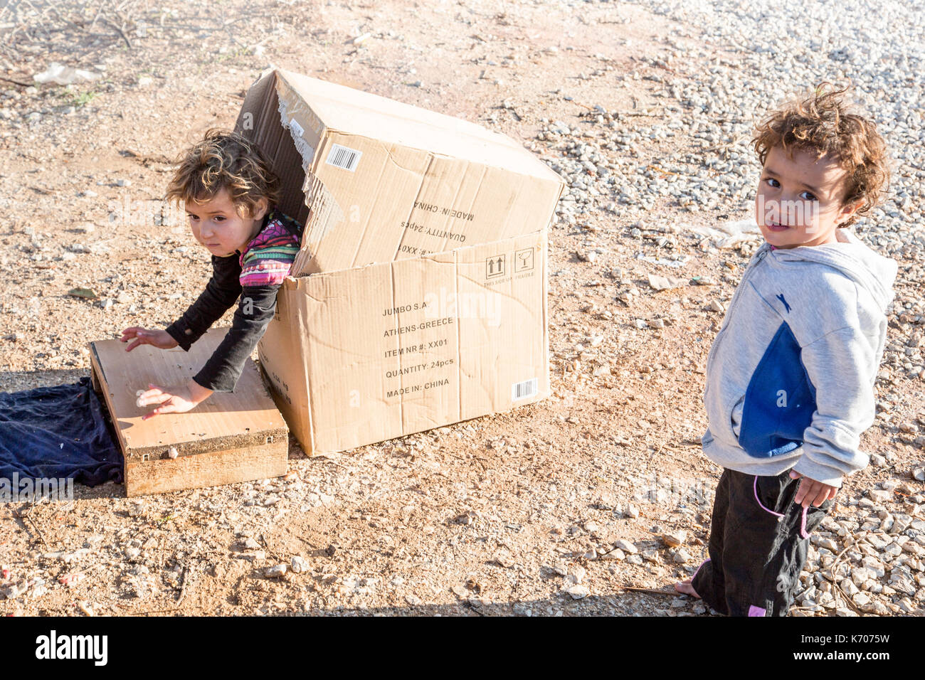 Un enfant joue gaiement dans une boîte en carton sur le sol pierreux de la ritsona camp de réfugiés en grèce comme son collègue regarde la caméra. Banque D'Images