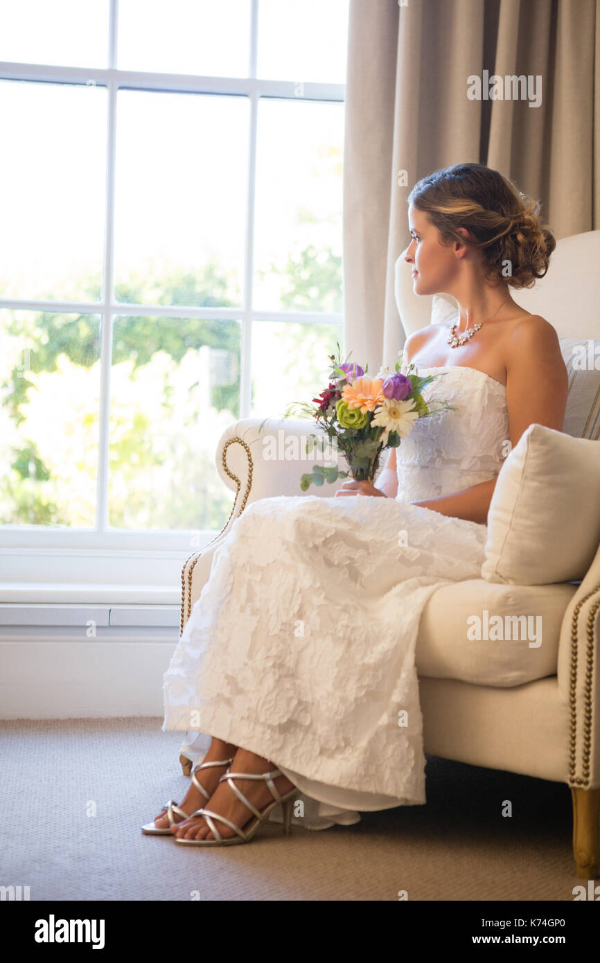 Belle bride holding bouquet à la fenêtre à travers tout en restant assis sur un fauteuil à la maison Banque D'Images