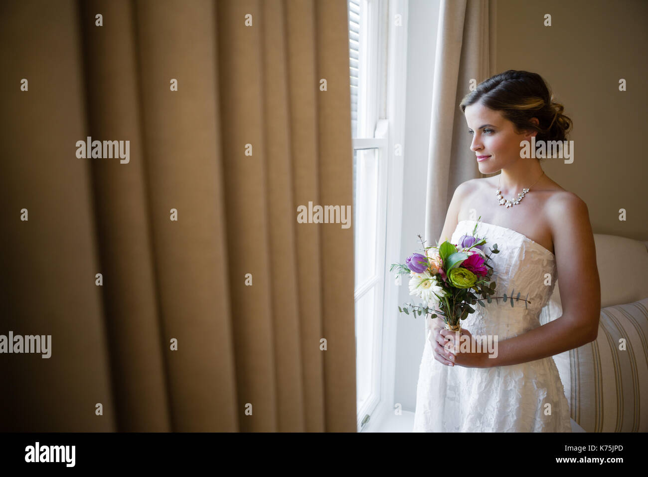 Smiling bride holding bouquet tout en regardant par la fenêtre à la maison Banque D'Images