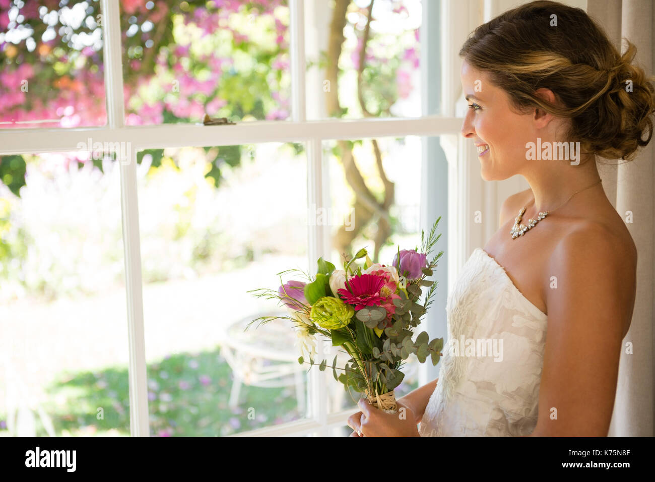 Vue latérale du smiling beautiful bride holding bouquet tout en regardant par la fenêtre à la maison Banque D'Images
