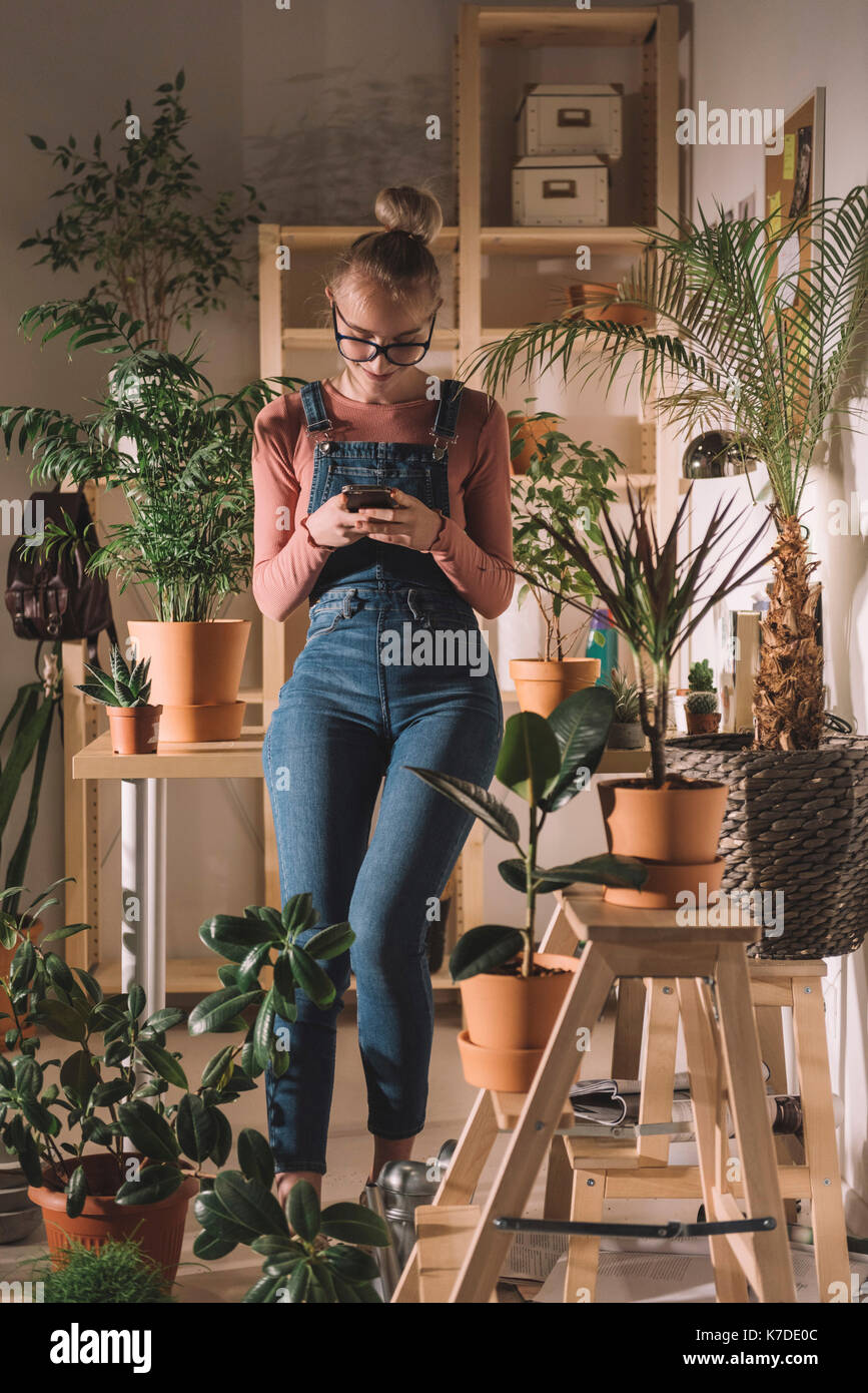 Woman using mobile phone tout en s'appuyant sur le tableau de plantes en pot à la maison Banque D'Images