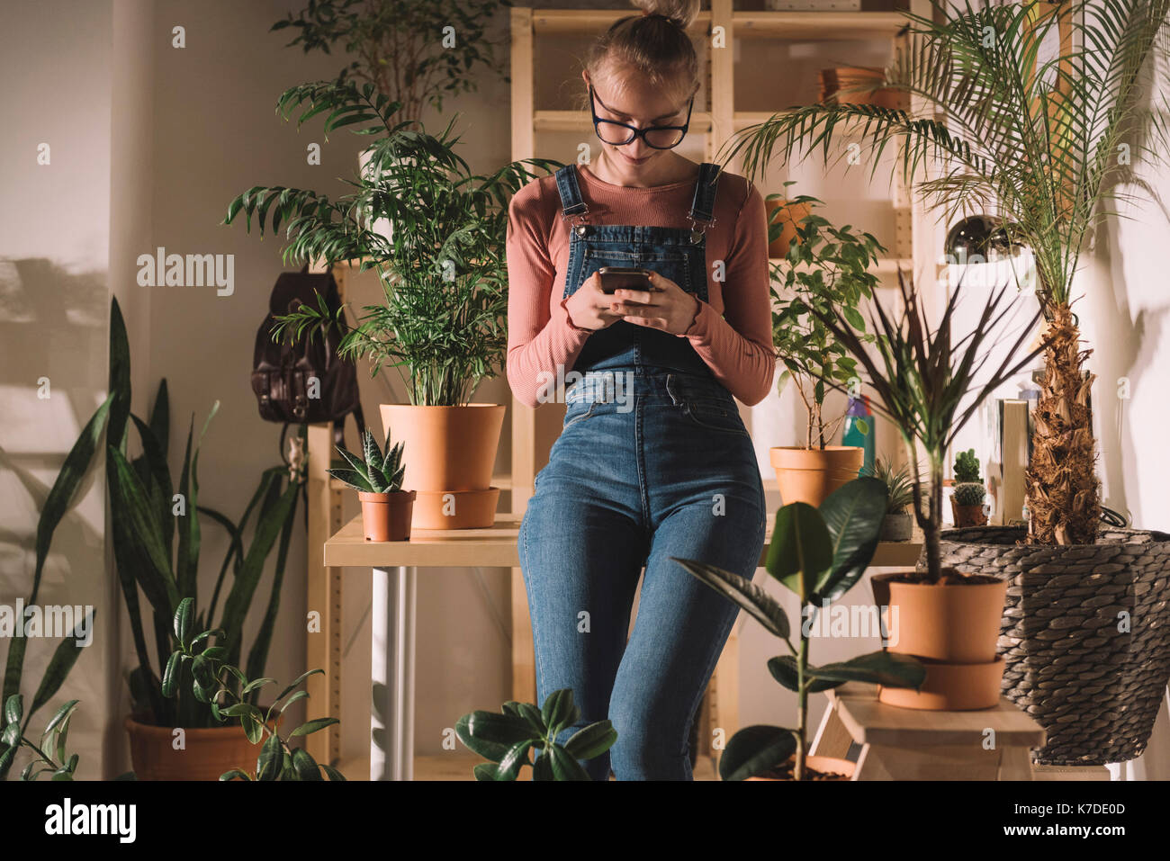 Femme à l'aide de smart phone tout en s'appuyant sur le tableau de plantes en pot à la maison Banque D'Images
