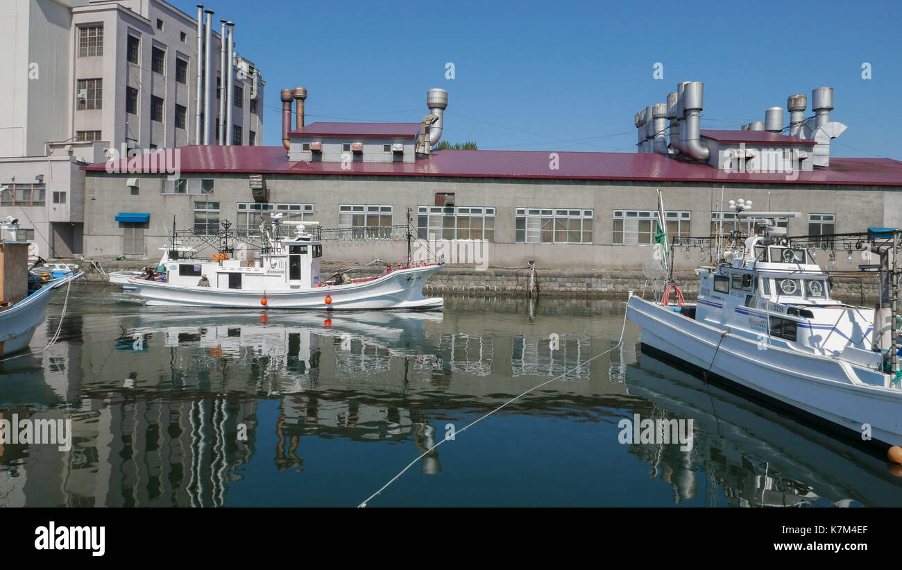 Coup horizontal de bateau de pêche blanche vers la mer d'Otaru canal avec usine en arrière-plan. Réflexions d'immeubles et de bateaux dans l'eau bleue Banque D'Images