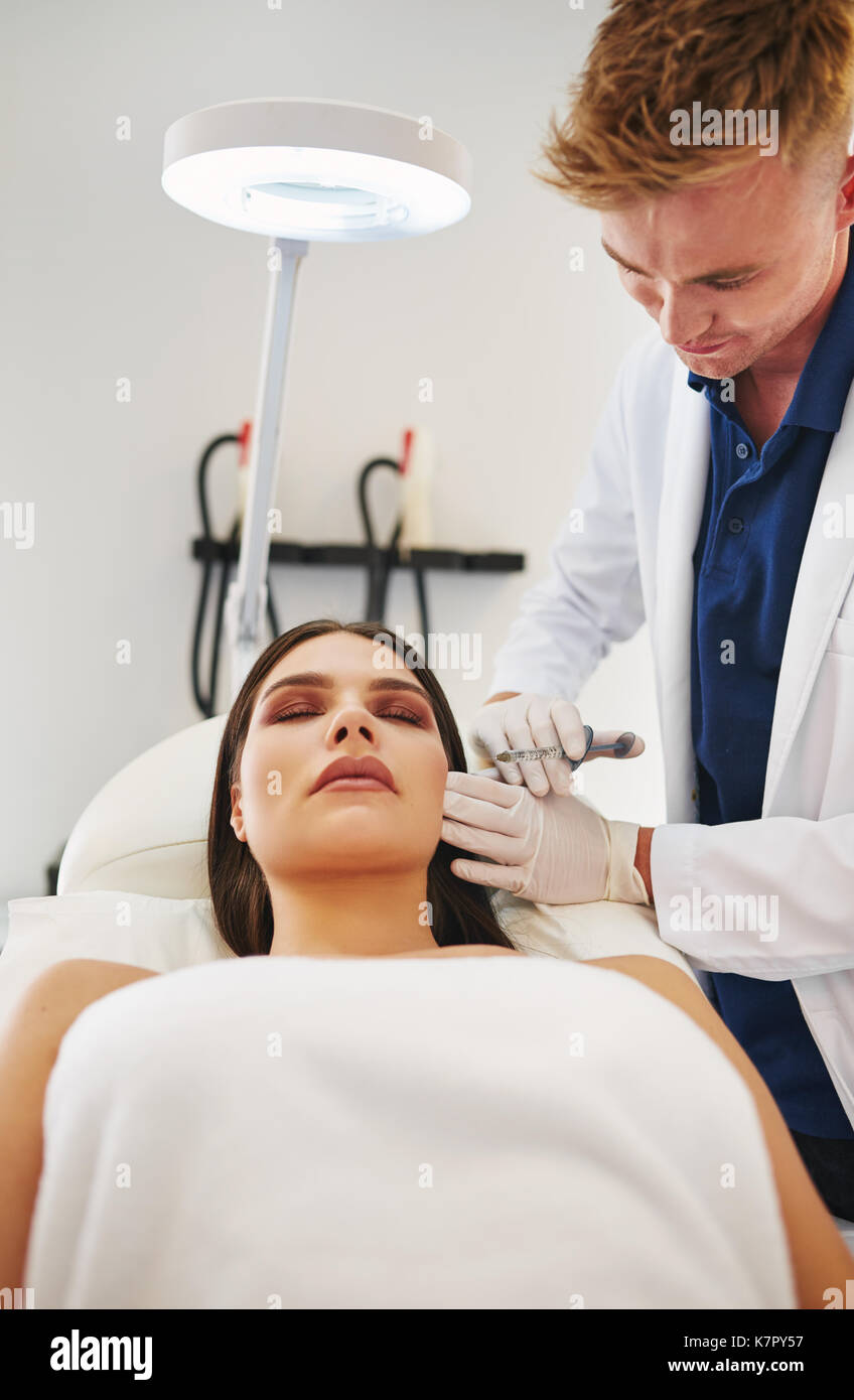 Jeune homme médecin d'effectuer des injections de botox à la joue d'une femme allongée sur une table dans une clinique de beauté Banque D'Images