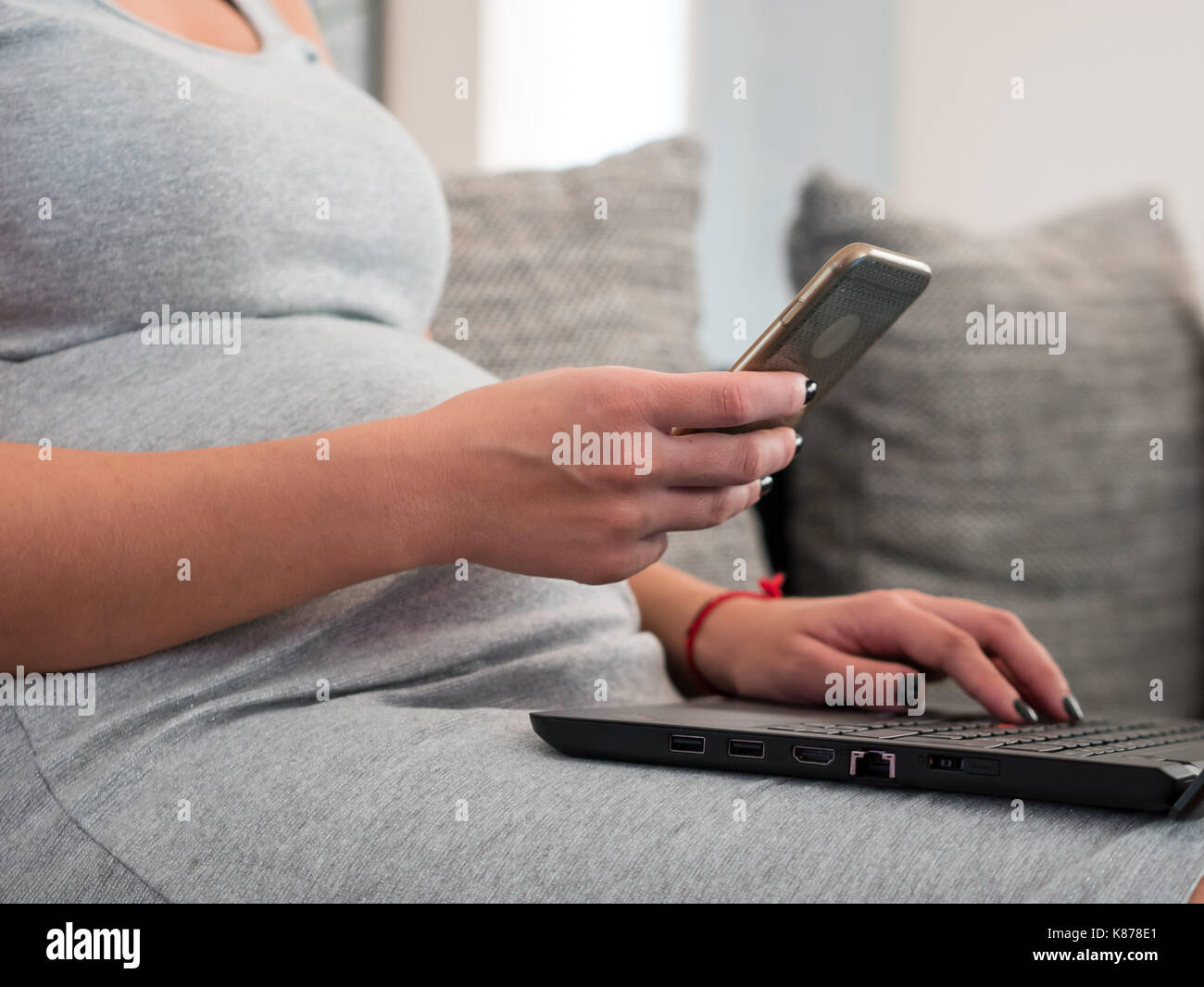 Pregnant woman sitting on sofa at home et à l'aide de l'ordinateur portable et smartphone pour les entreprises Banque D'Images