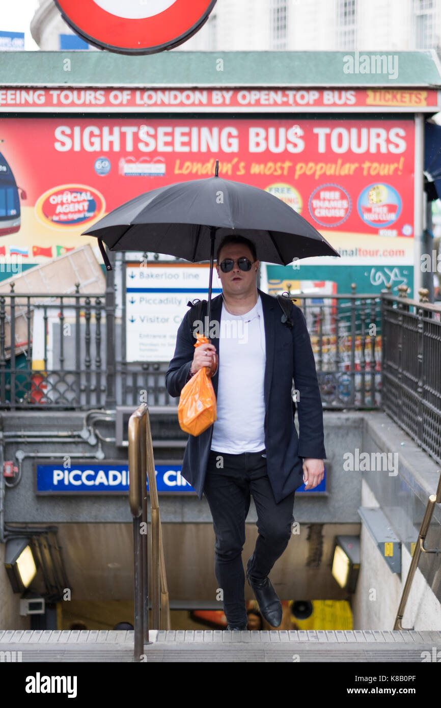 Homme de couleur blanche portant des lunettes de soleil émerge d'une station de métro de Londres avec son parapluie ouvert au cours d'une douche à effet pluie. Banque D'Images