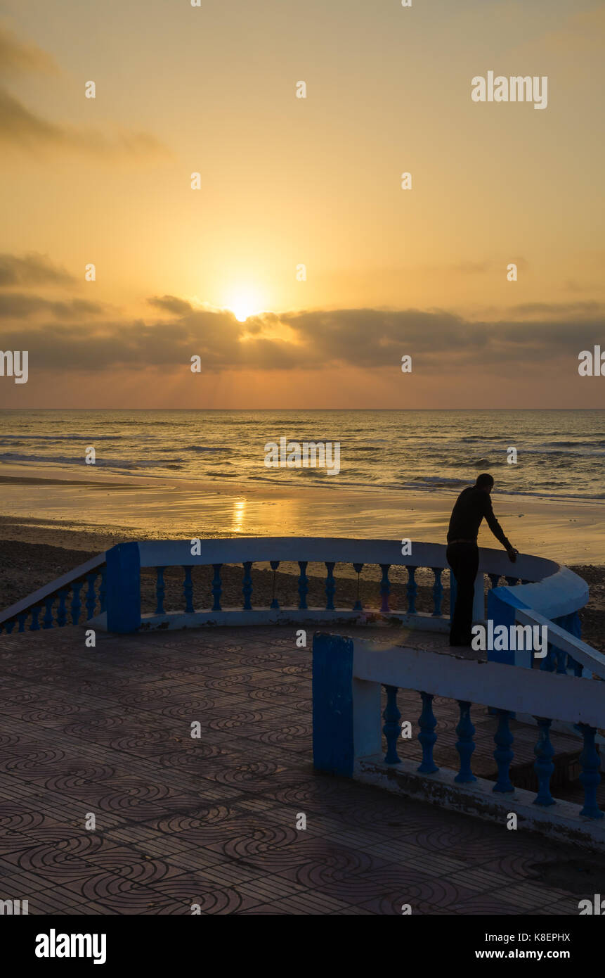 Silhouette of man standing at beach promenade avec passage de pierre pendant le coucher du soleil à Sidi Ifni, Maroc, afrique du nord. Banque D'Images