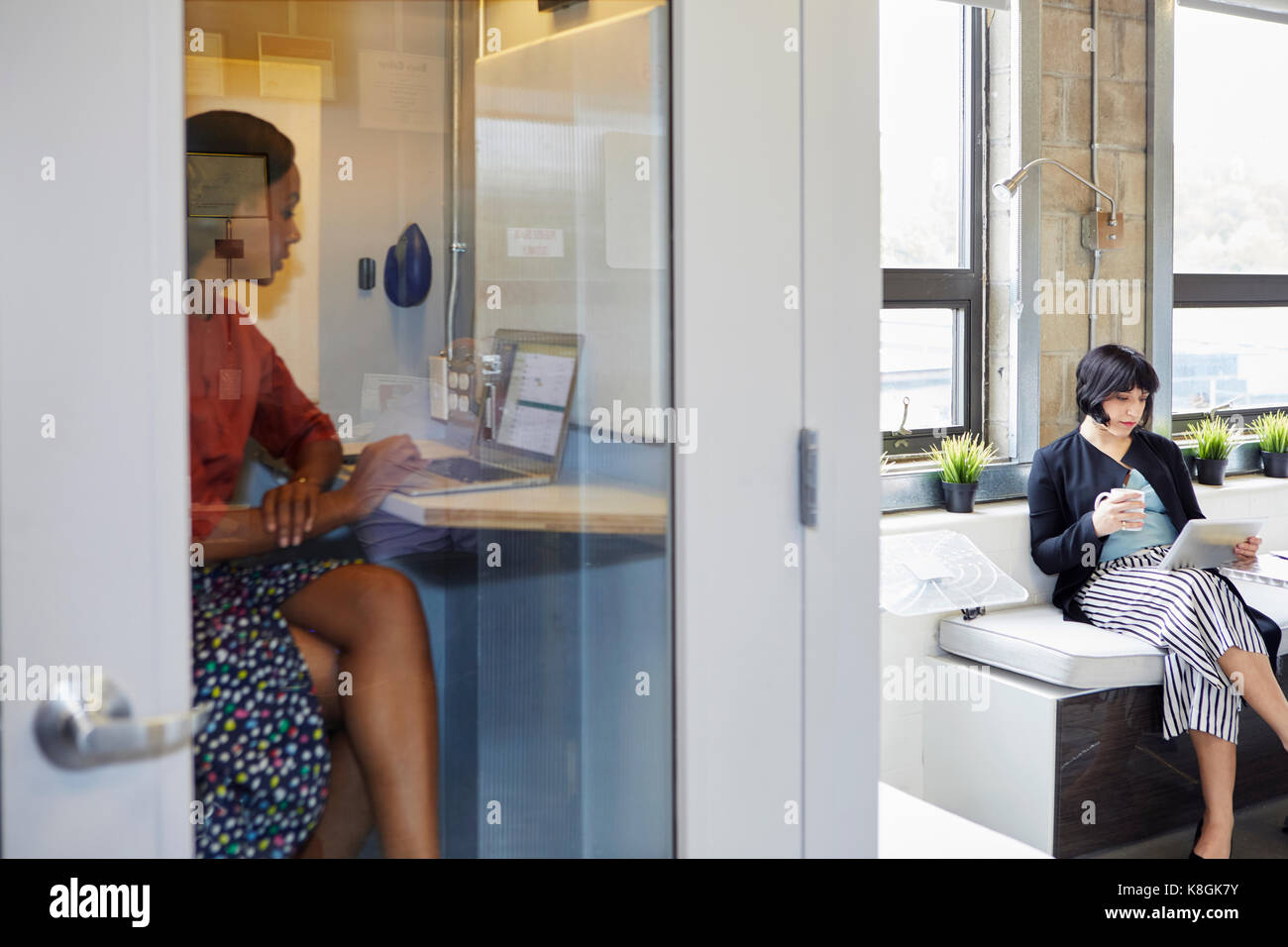 Jeune femme dans une cabine téléphonique de bureau, à l'aide d'un ordinateur portable, collègue assis à l'extérieur Banque D'Images