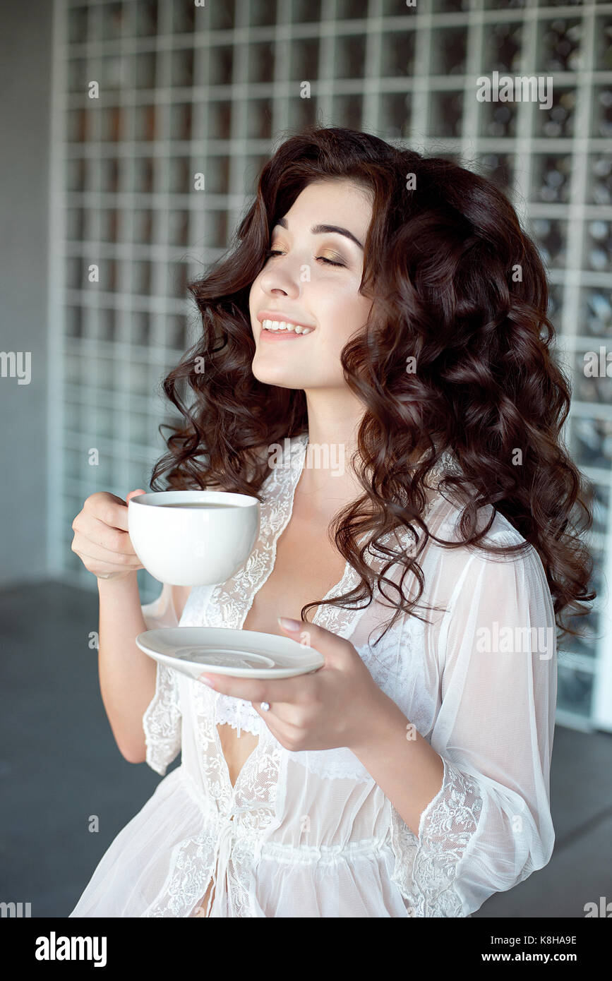 Attractive smiling girl savourer une tasse de café dans un peignoir blanc  transparent. modèle avec les cheveux bouclés brun et beau corps. tourné  mi-longueur Photo Stock - Alamy