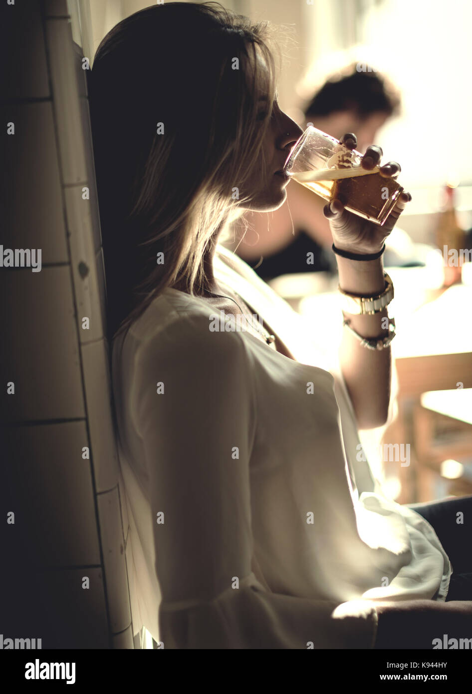 Vue latérale d'une femme avec de longs cheveux blonds debout à l'intérieur, boire de la bière dans un verre. Banque D'Images