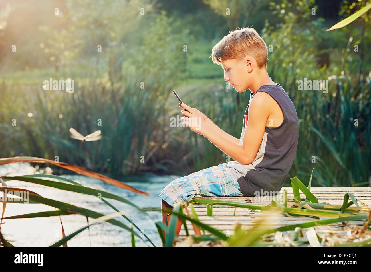 L'été à la campagne. garçon assis sur la jetée du lac. petit photographe prendre photo de la libellule par smart phone appareil photo. Banque D'Images