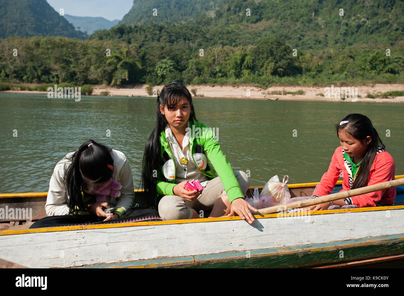 Trois adolescentes une holding a mobile phone s'asseoir dans une rivière canoë avec les montagnes derrière à Muang Ngoi Laos Banque D'Images