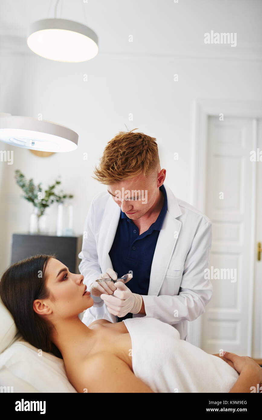 Médecin homme d'effectuer des injections de botox pour les lèvres d'une jeune femme brune allongé sur une table dans une clinique de beauté Banque D'Images