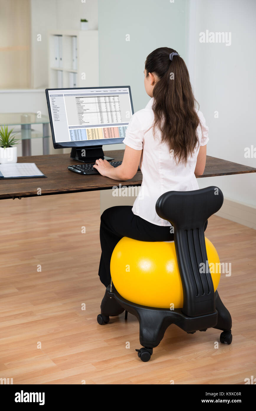 Young businesswoman sitting on fitness ball tout en travaillant sur computer in office Banque D'Images