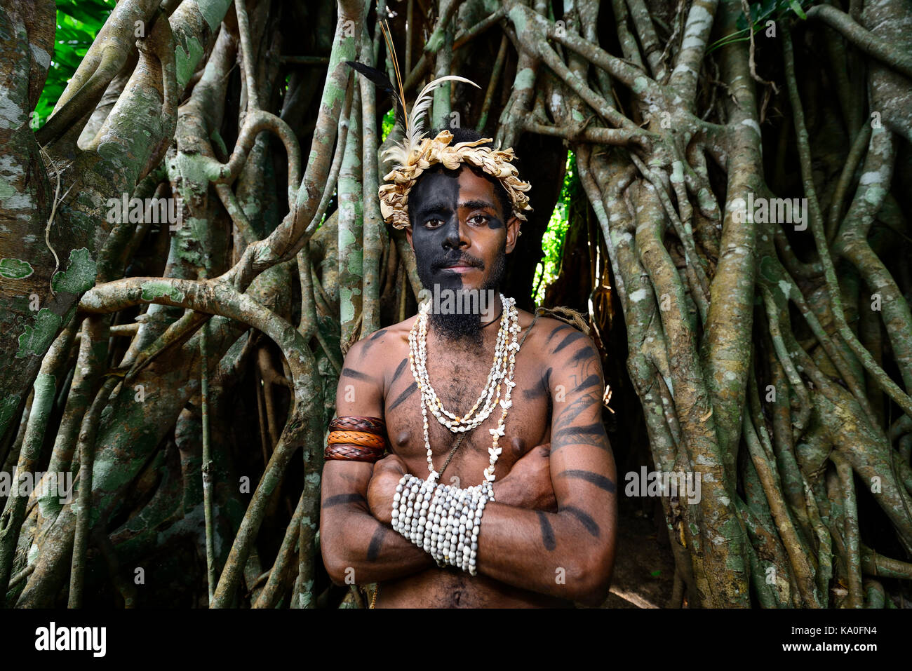 Guerrier Tribal devant les racines de l'arbre de banian (Ficus benghalensis), ekasup village culturel, l'île d'Efate, Vanuatu Banque D'Images