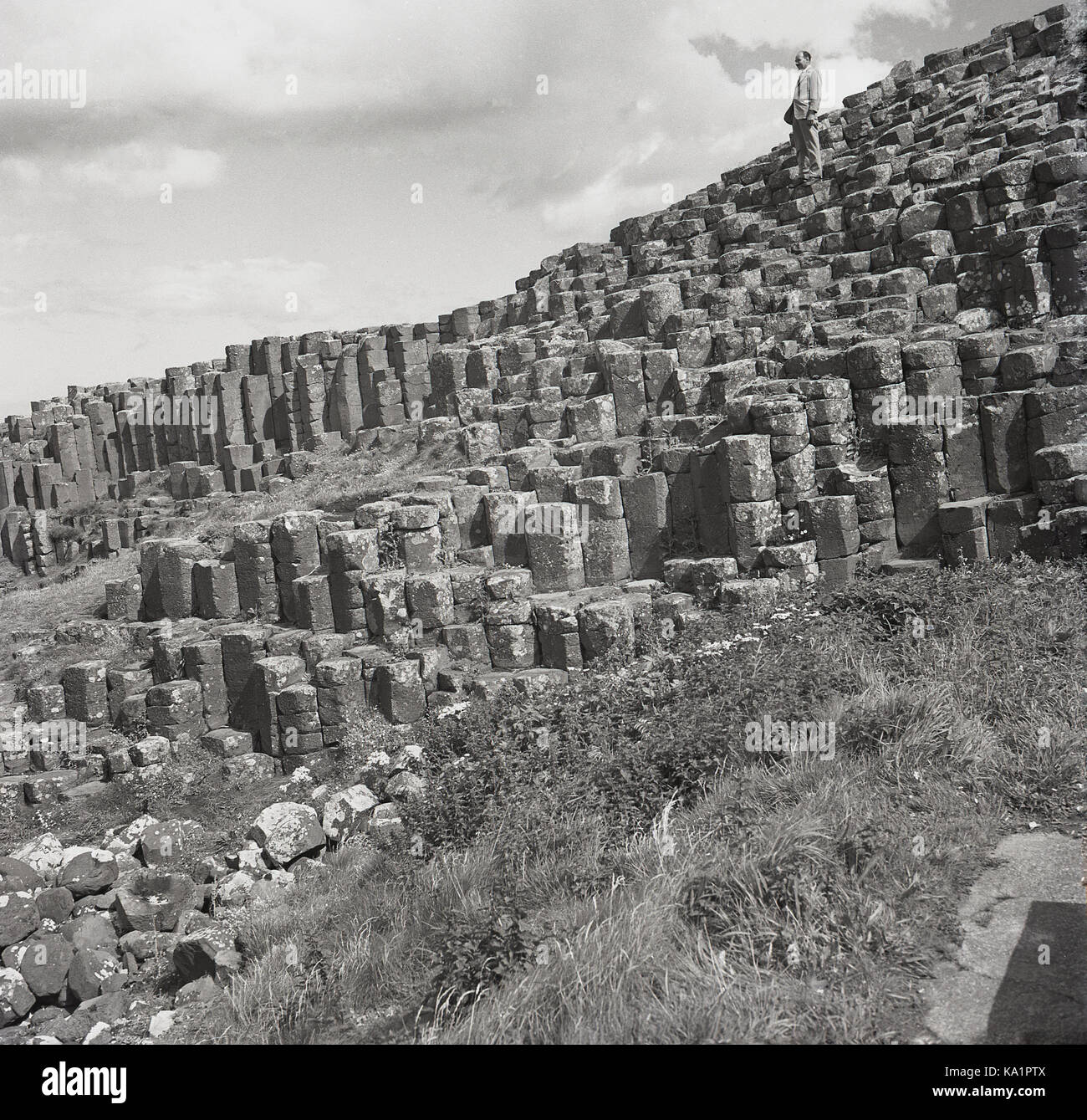 Années 1950, historique, un gentleman se tenant au-dessus des anciennes formations rocheuses volcaniques de colonnes de basalte imbriquées, à la célèbre chaussée des géants, sur la côte de l'Atlantique Nord, près de Bushmills, Co Antrim, Irlande du Nord, Royaume-Uni. Banque D'Images