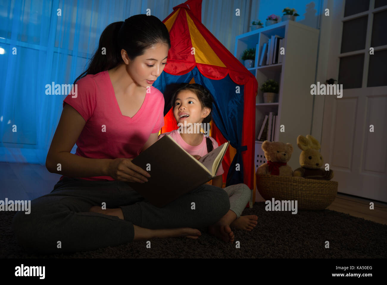 Slim élégance mère tenant livre parlant histoire avant de smiling happy little girl et assis en face d'enfants toy tente de nuit sur Banque D'Images