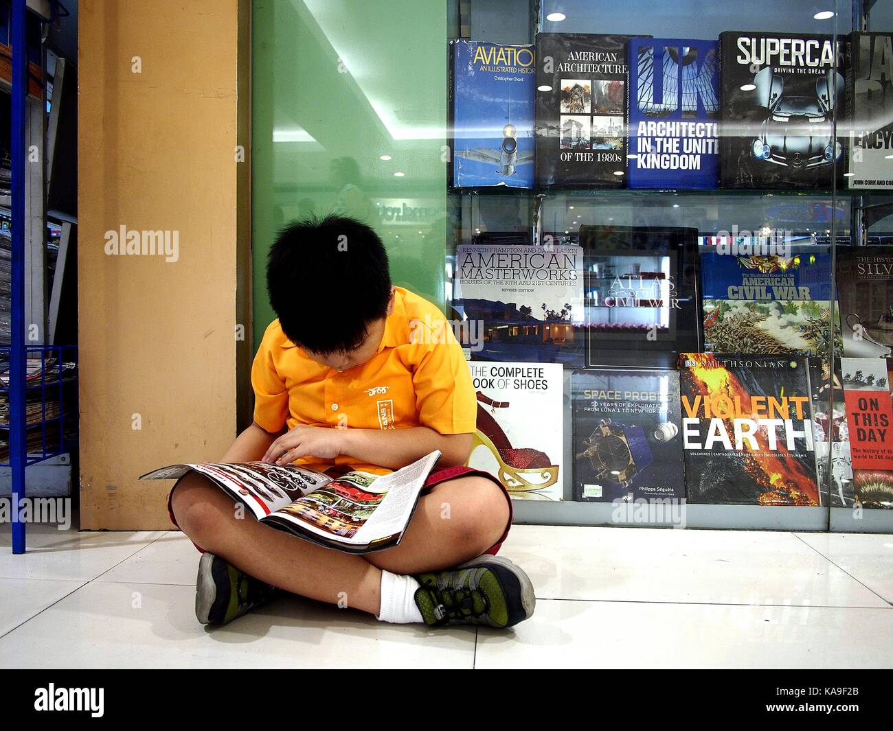 Mandaluyong City, Philippines - 24 septembre 2017 : un jeune garçon est assis sur le sol et lit un magazine à l'extérieur d'un employé et l'excédent de librairie. Banque D'Images