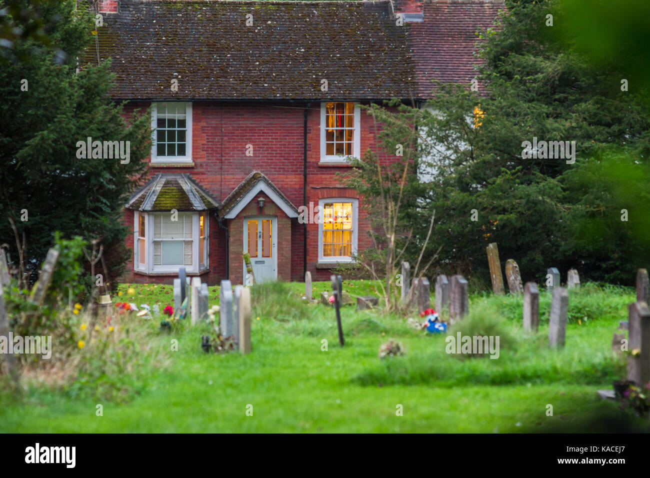 Vue sur le cimetière de l'église All Saints avec un cottage en brique rouge avec la lumière ou dans l'arrière-plan, à l'Est Meon, Hampshire, Royaume-Uni Banque D'Images