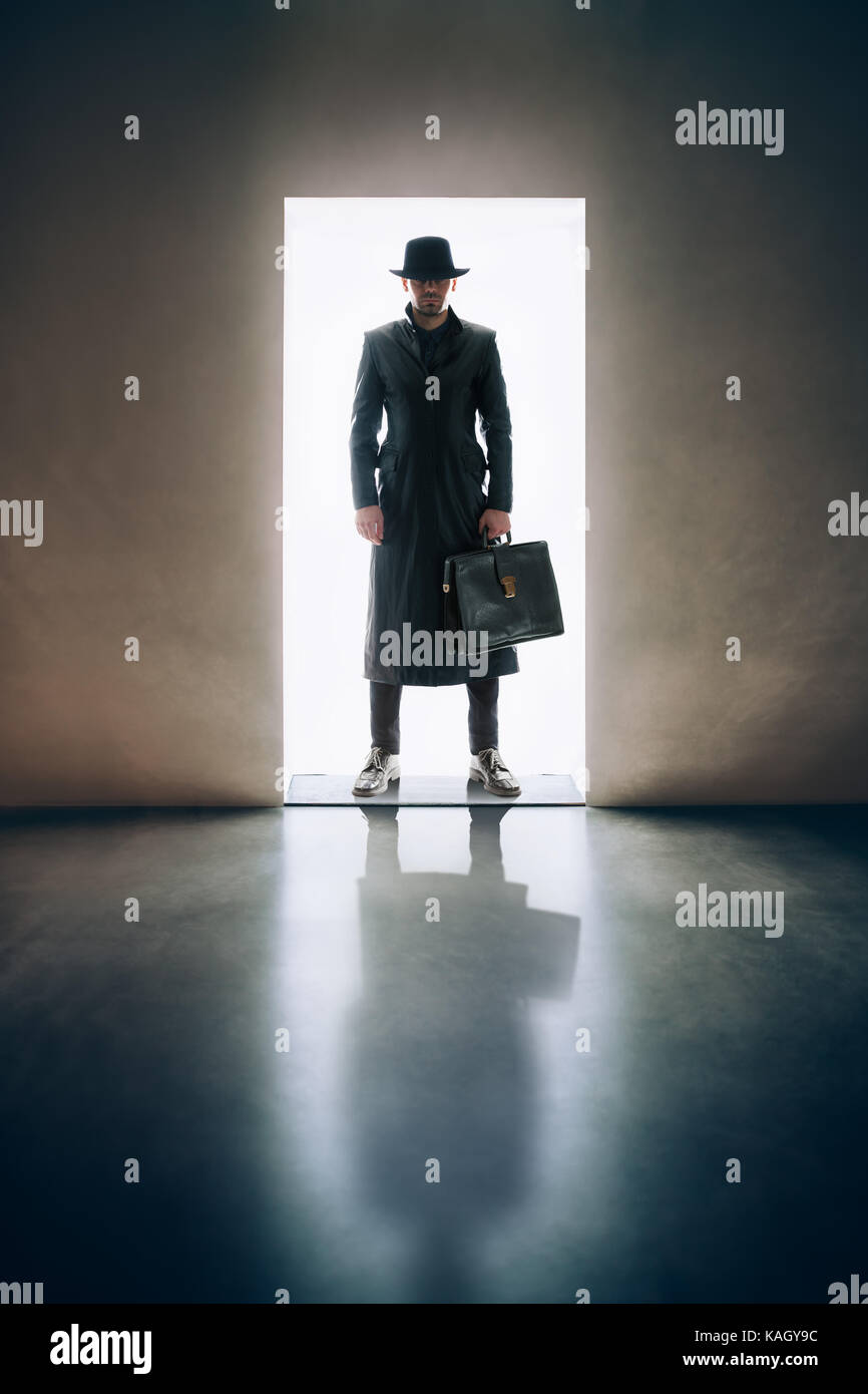 L'homme silhouette en imperméable et chapeau debout dans la lumière de l'ouverture de la porte dans une pièce sombre Banque D'Images