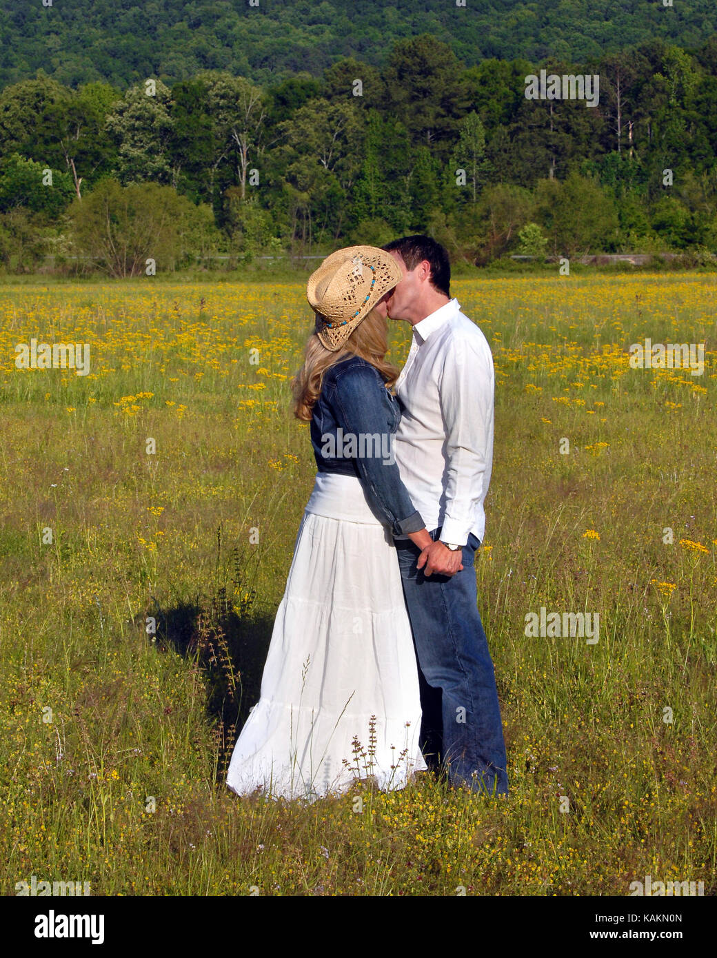 Jeune couple embrasser dans un champ de fleurs jaunes. tenant les mains ils  baiser. Elle porte un chapeau de cowboy et denim veste. Il porte des jeans  et w Photo Stock -