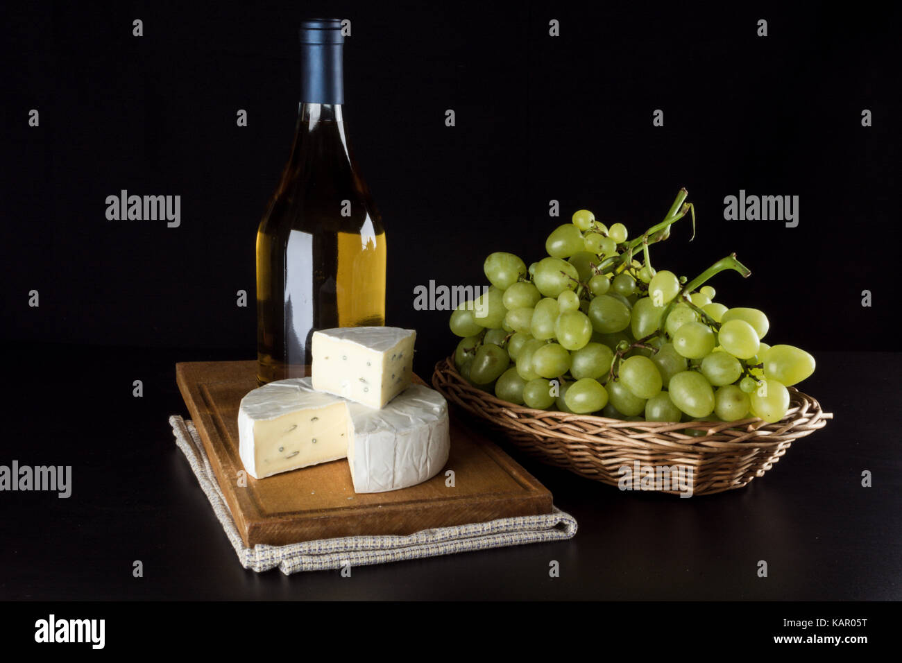 Une bouteille de vin blanc à l'arrière-plan, des raisins dans un panier et dor le fromage bleu sur fond noir Banque D'Images