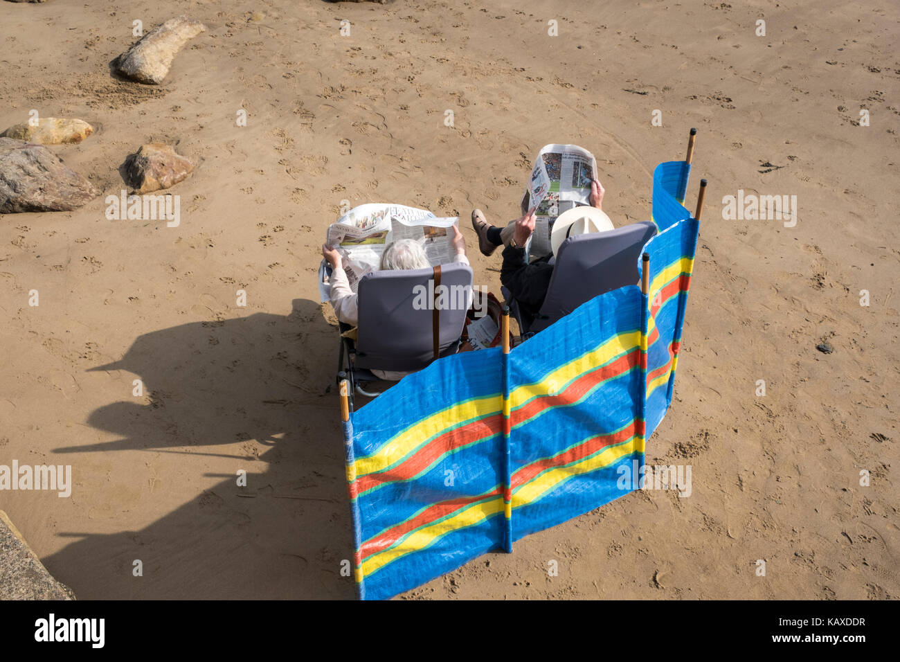 Couple de personnes âgées lire les journaux derrière un brise-vent sur une plage de sable, à l'ensoleillement, Scarborough Banque D'Images
