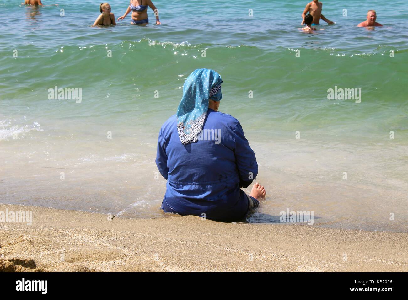 Juillet, 2017 - une vieille femme musulmane est assis sur le sable dans une zone de déferlement sur l'arrière-plan de touristes de l'Europe (Istanbul, Turquie). Banque D'Images