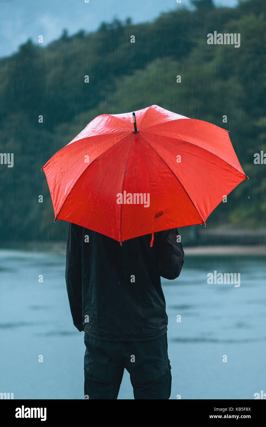 Homme avec parapluie rouge prévoit de la pluie en face d'un lac. triste et solitaire à la personne de sexe masculin dans la distance. Banque D'Images
