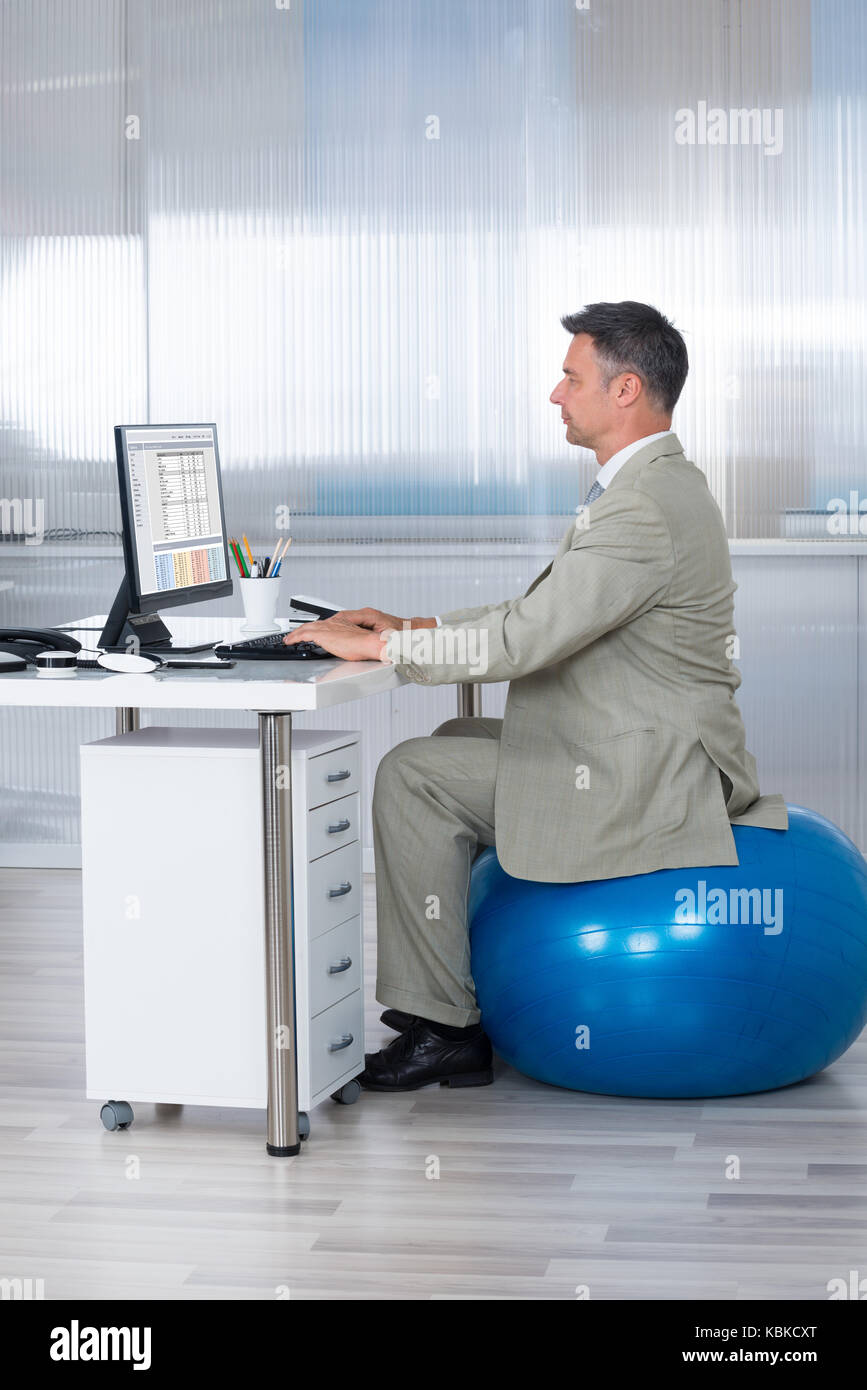 Vue latérale du businessman using computer while sitting on exercise ball at office Banque D'Images