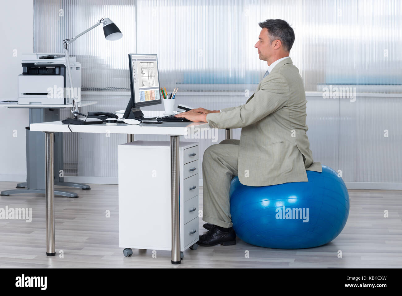Vue latérale du businessman using computer while sitting on exercise ball at office Banque D'Images