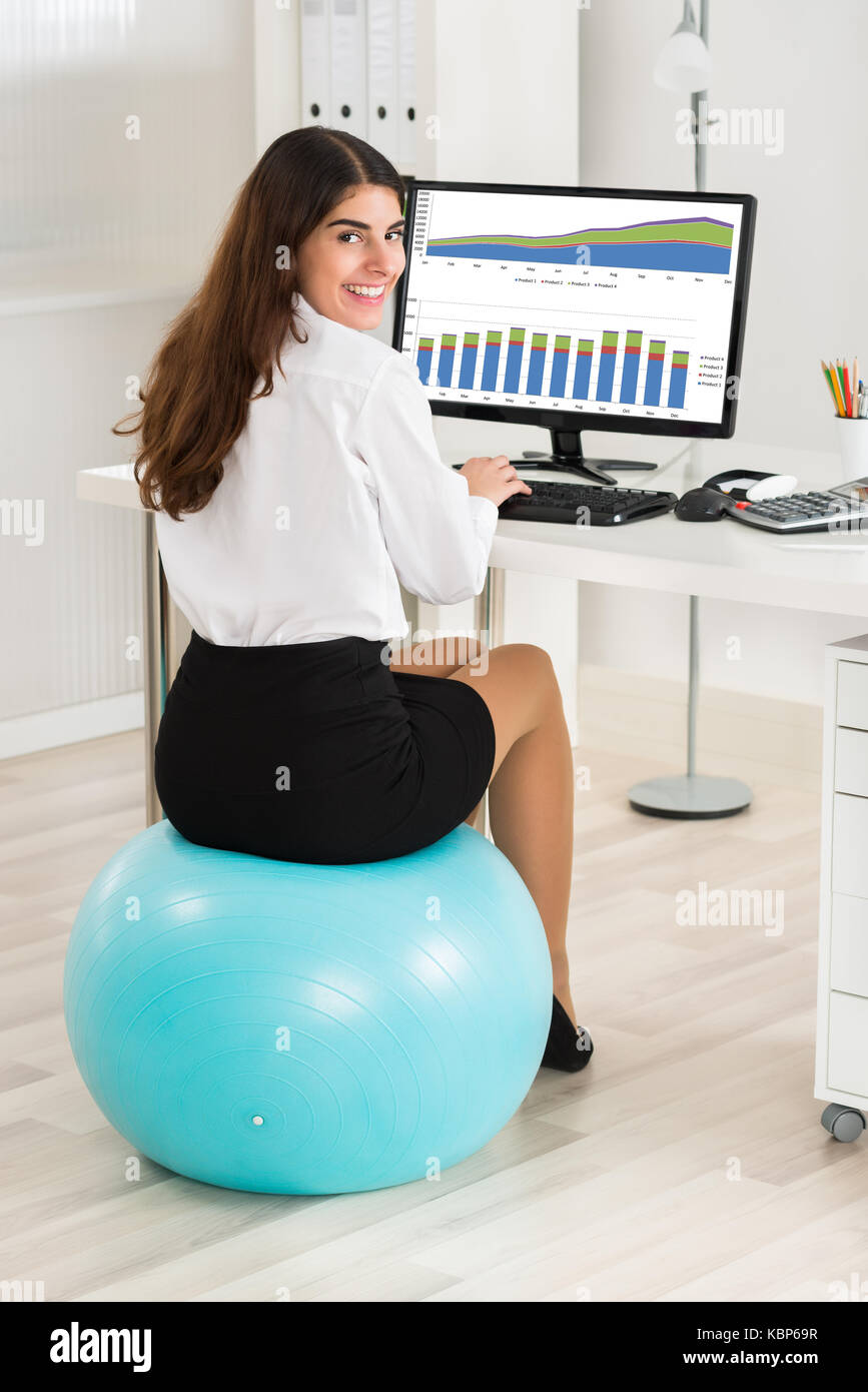 Portrait of happy young businesswoman using computer while sitting on exercise ball in office Banque D'Images