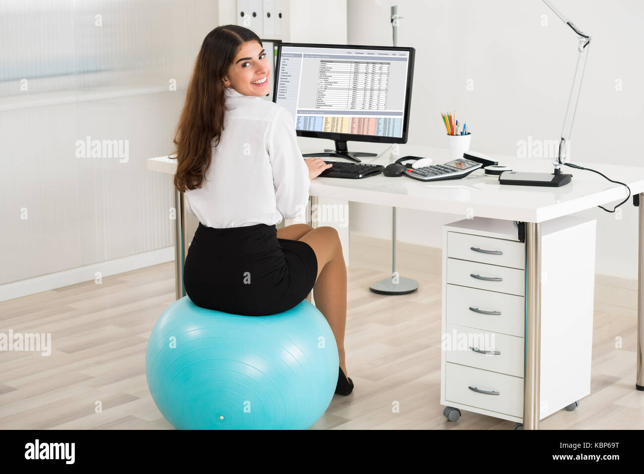 Portrait of happy young businesswoman using computer while sitting on exercise ball in office Banque D'Images