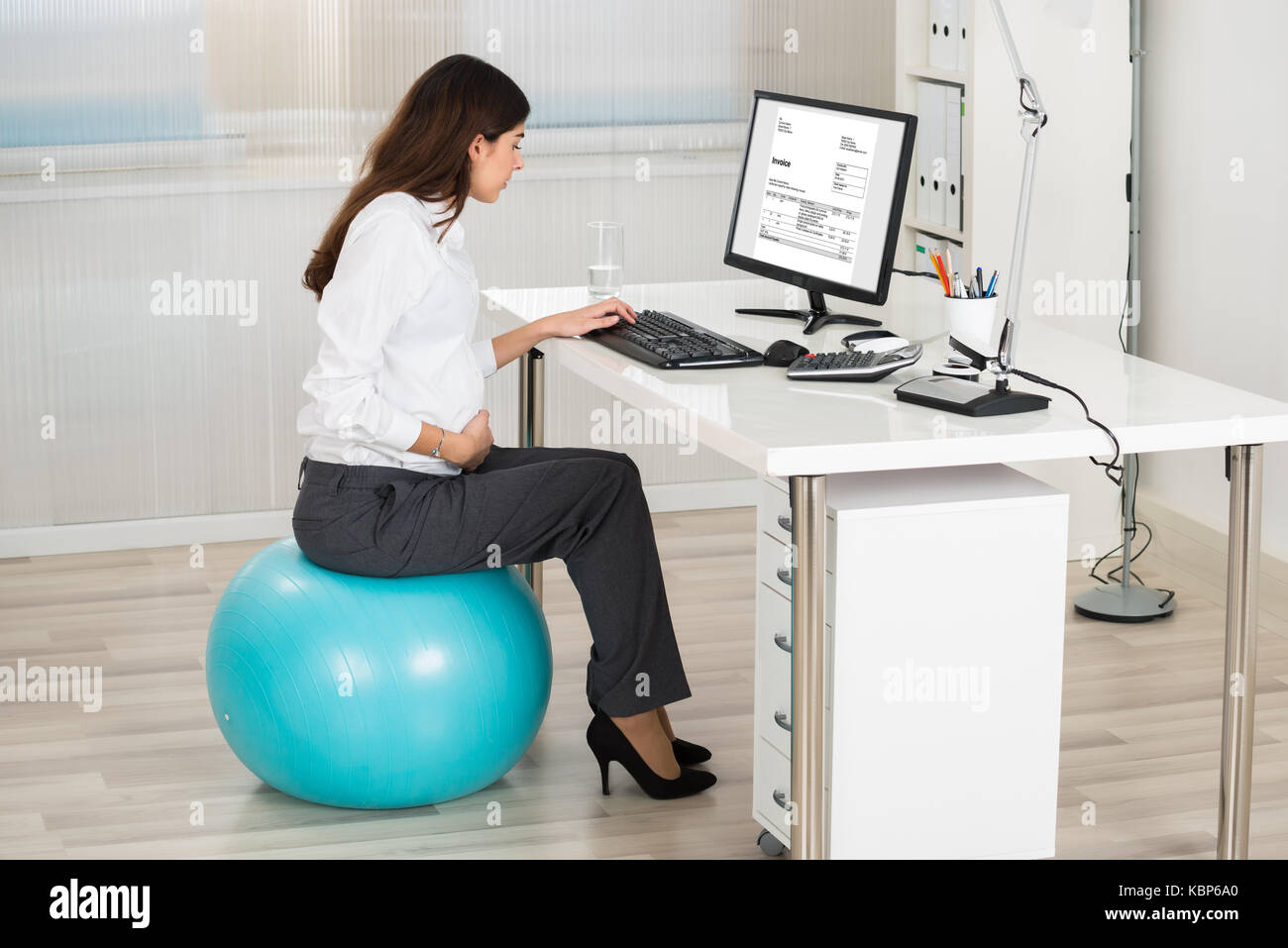 Vue du côté des femmes enceintes businesswoman using computer while sitting on exercise ball in office Banque D'Images