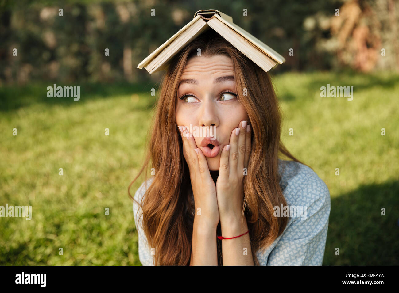 Funny brunette woman sitting in park avec livre sur sa tête en tenant les bras sur les joues et à la suite Banque D'Images