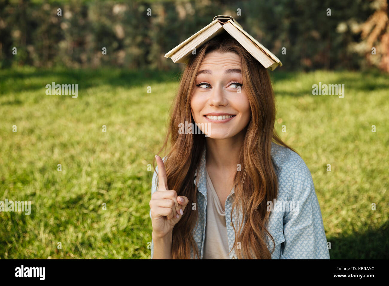 Smiling brunette woman sitting in park avec livre sur sa tête tout en regardant ailleurs Banque D'Images