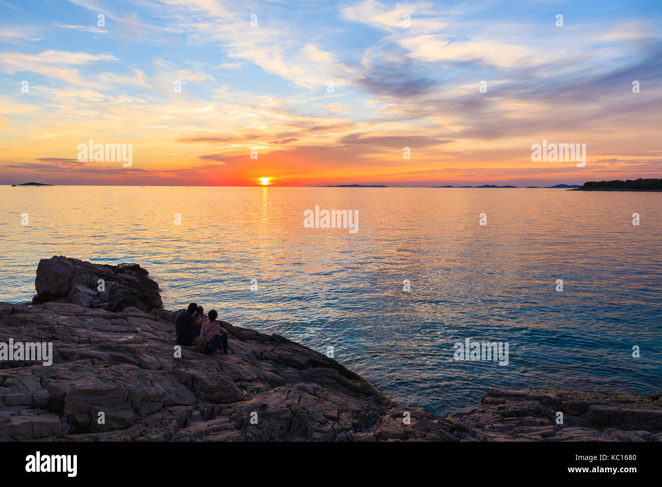 Couple non identifié de personnes assises sur les rochers et regardant le coucher du soleil dans la ville de Primosten, Croatie, Dalmatie Banque D'Images