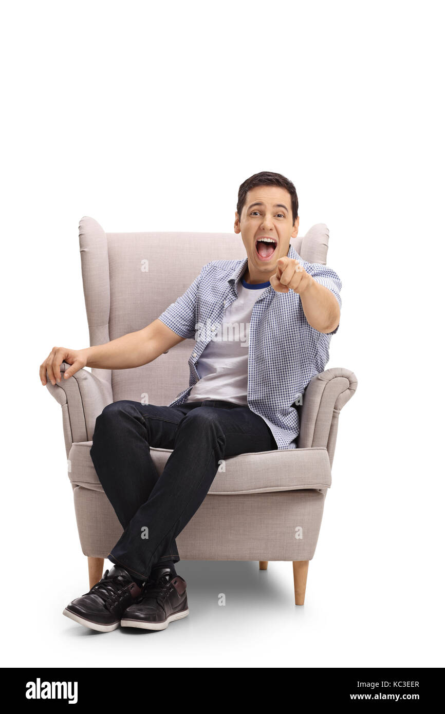 Jeune homme assis dans un fauteuil, pointant sur l'appareil photo et rire isolé sur fond blanc Banque D'Images