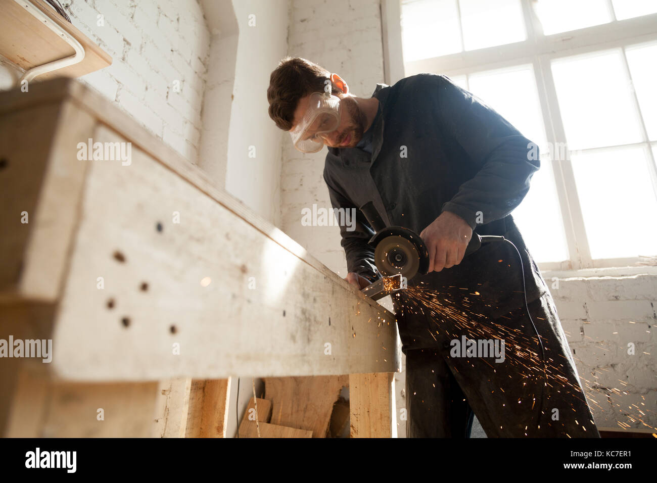Travailleur dans des lunettes de travailler en petit atelier intérieur à l'aide de la meuleuse d'angle de barre pour couper l'acier, jetant des étincelles de métal mac Banque D'Images