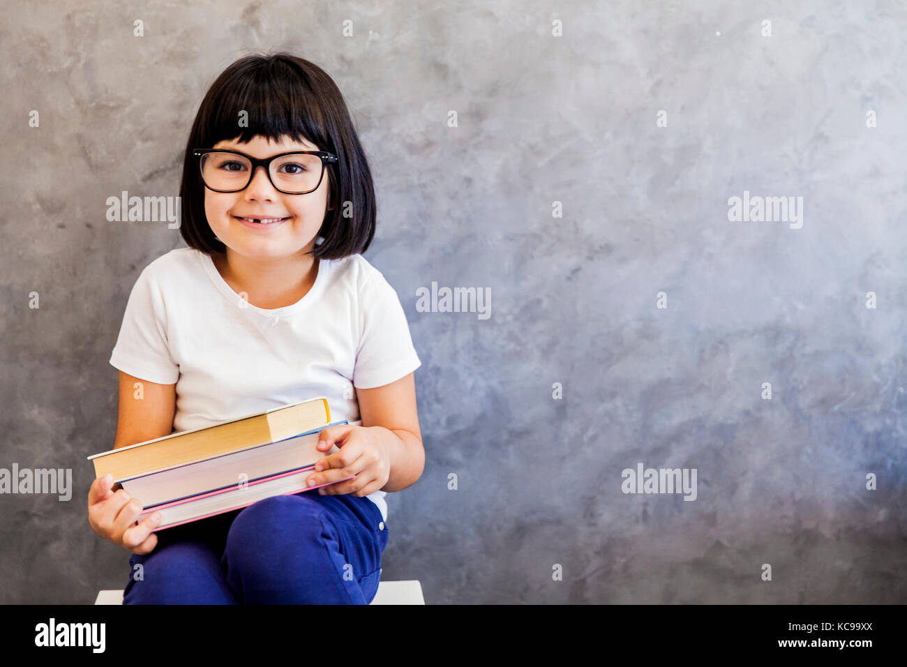 Jolie petite fille aux cheveux noir verres holding books par le mur Banque D'Images