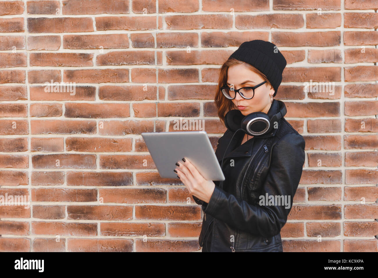 Taille portrait of young woman standing dans la rue à côté du mur de brique, écouter de la musique en ligne avec les écouteurs de tablette. Heureux belle femme usin Banque D'Images