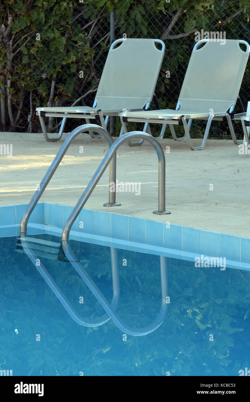 Deux chaises longues à côté d'une piscine et reflets dans l'eau bleu clair avec quelques mains courantes et les étapes pour entrer l'eau. Banque D'Images