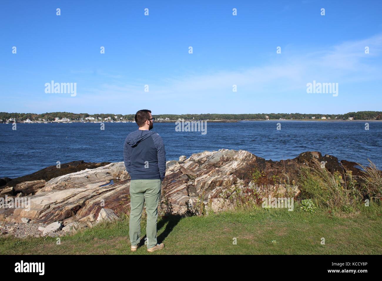 Homme debout près d'un rocher sur l'herbe avec un ciel clair et de l'océan en arrière-plan Banque D'Images