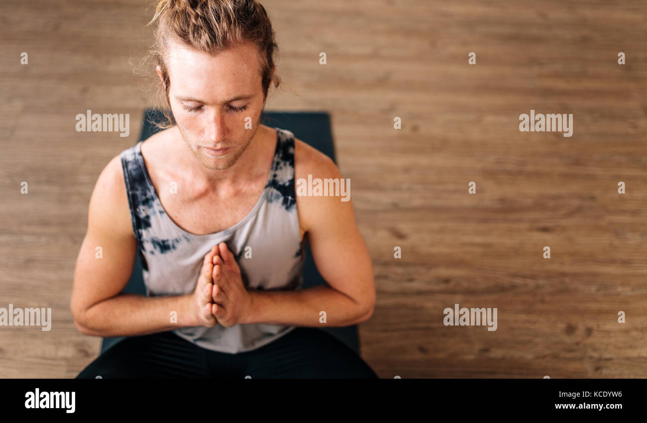 Vue de tête de jeune homme en forme pratique yoga assis sur le sol avec ses mains jointes. gars de fitness faisant de la méditation yoga à l'intérieur. Banque D'Images