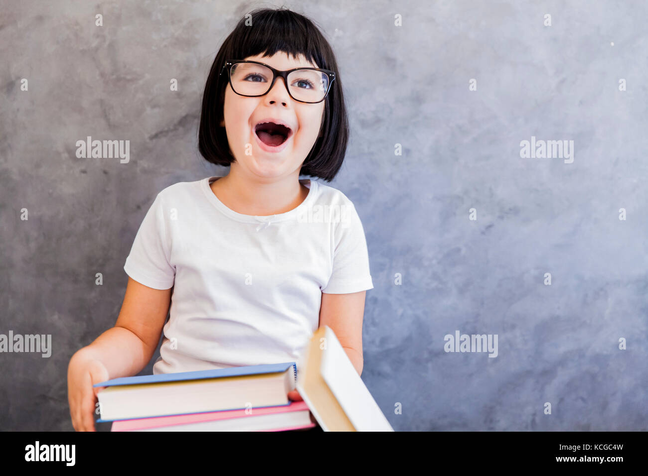 Jolie petite fille aux cheveux noir verres holding books par le mur Banque D'Images