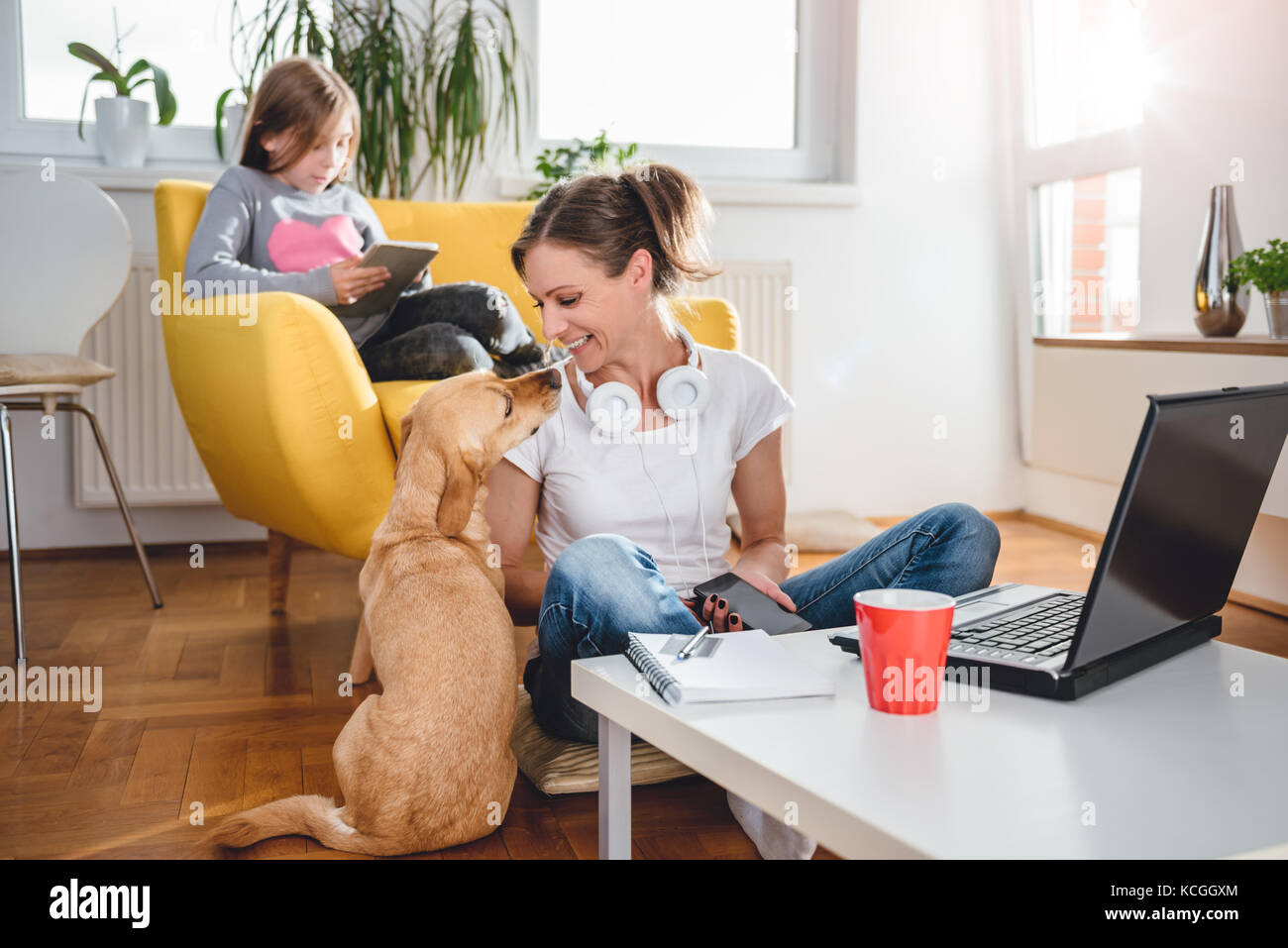 Femme assise sur le sol et caresser chien à la maison tandis que sa fille assise sur le fauteuil et l'utilisation de tablet Banque D'Images Femme assise sur le sol et caresser chien à la maison tandis que sa fille assise sur le fauteuil et l'utilisation de tablet Banque D'Images