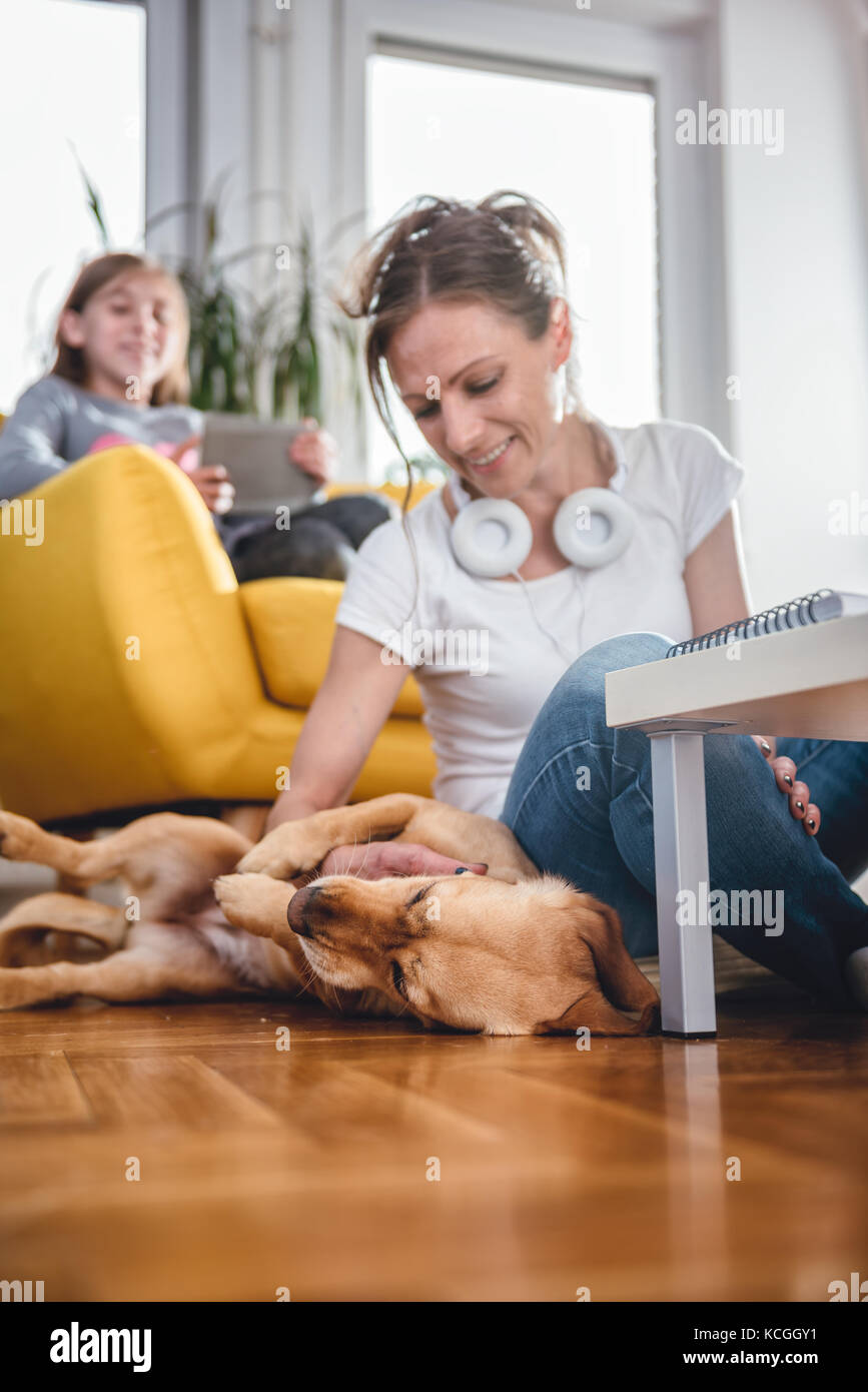 Femme assise sur le sol et caresser chien à la maison tandis que sa fille assise sur le fauteuil et l'utilisation de tablet Banque D'Images Femme assise sur le sol et caresser chien à la maison tandis que sa fille assise sur le fauteuil et l'utilisation de tablet Banque D'Images