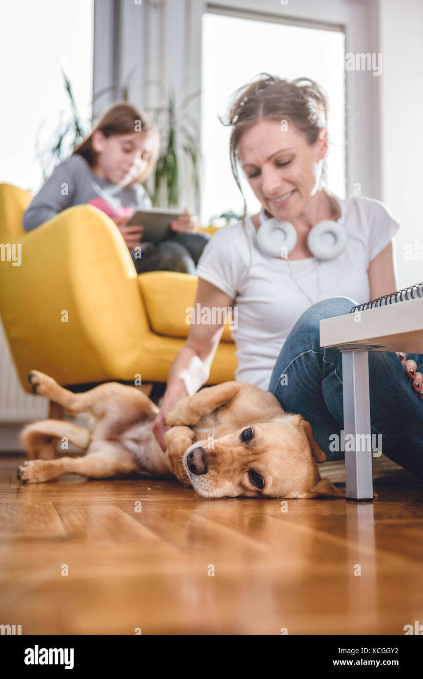 Femme assise sur le sol et caresser chien à la maison tandis que sa fille assise sur le fauteuil et l'utilisation de tablet Banque D'Images Femme assise sur le sol et caresser chien à la maison tandis que sa fille assise sur le fauteuil et l'utilisation de tablet Banque D'Images