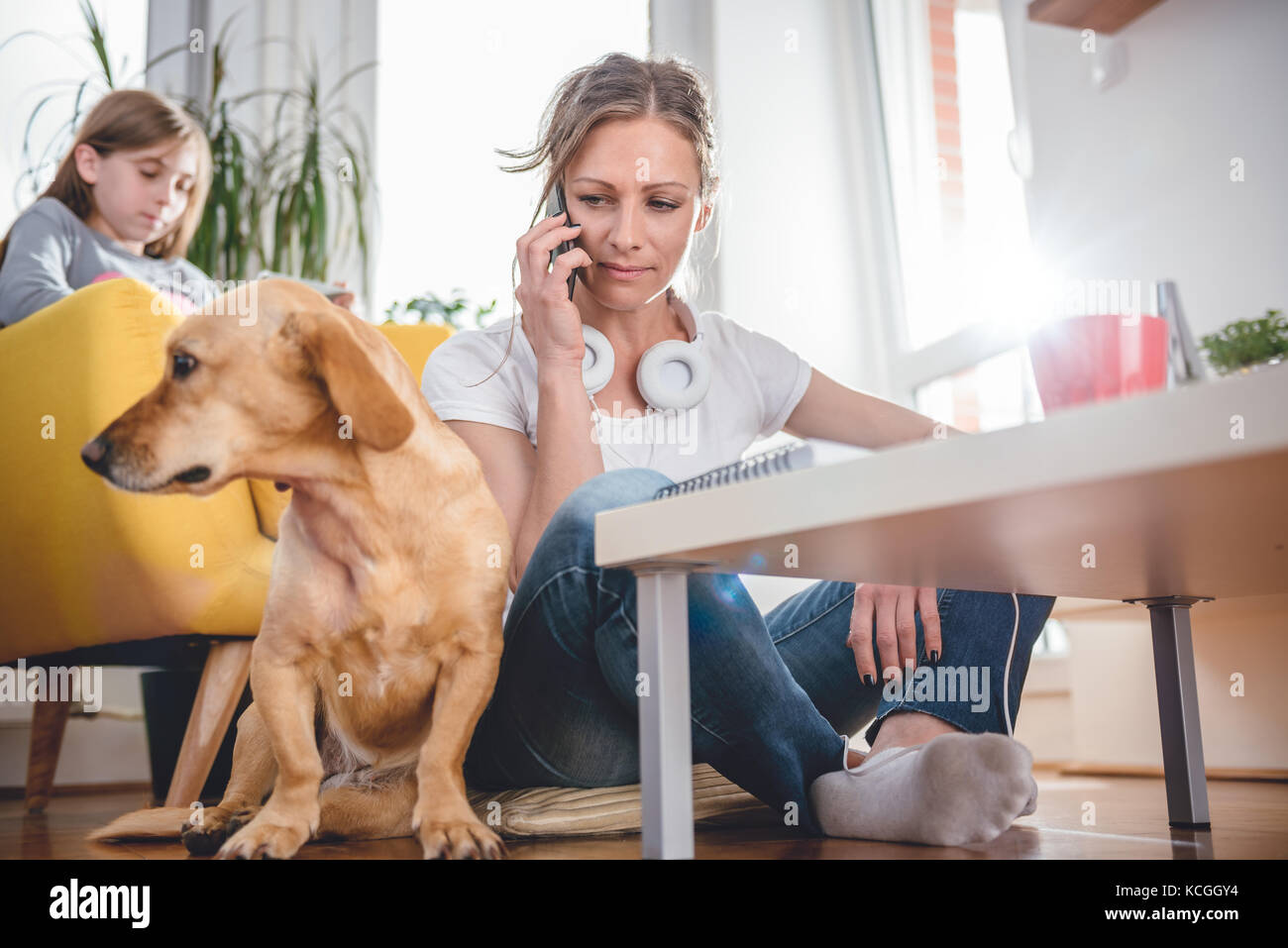 Femme assise sur le sol par le petit chien jaune et parler sur smart phone at home alors que fille assis dans le fauteuil jaune à l'arrière-plan Banque D'Images Femme assise sur le sol par le petit chien jaune et parler sur smart phone at home alors que fille assis dans le fauteuil jaune à l'arrière-plan Banque D'Images